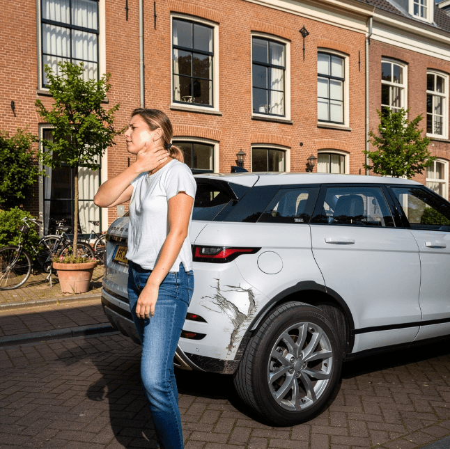 Persoon staat naast een stilstaande auto met beschadigingen aan de achterkant. Zij heeft pijn aan haar nek.