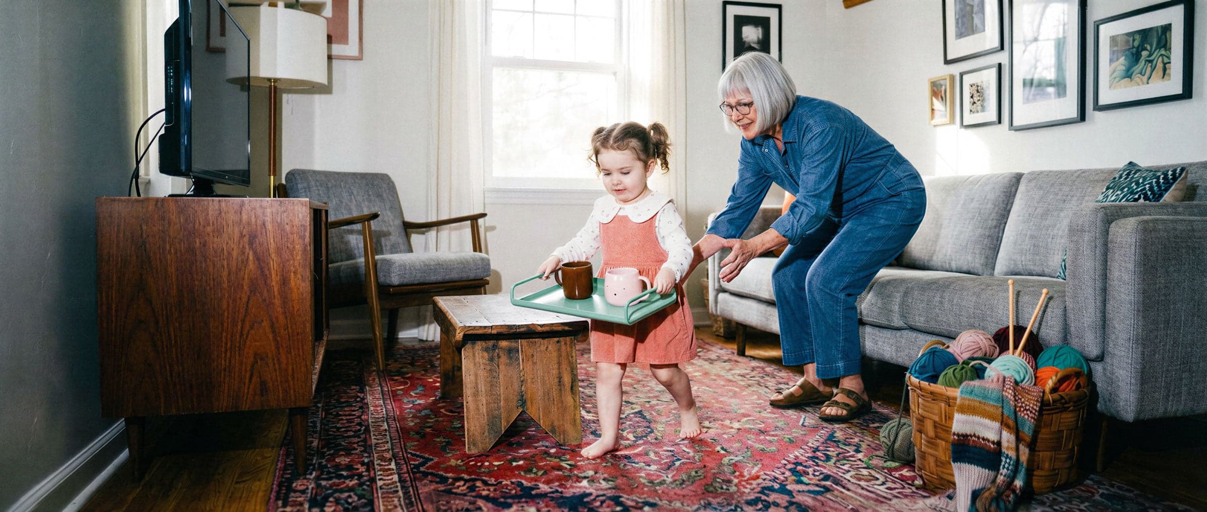 Jong kind loopt met een dienblad met speelgoedkopjes door de woonkamer, terwijl een grootouder meekijkt
