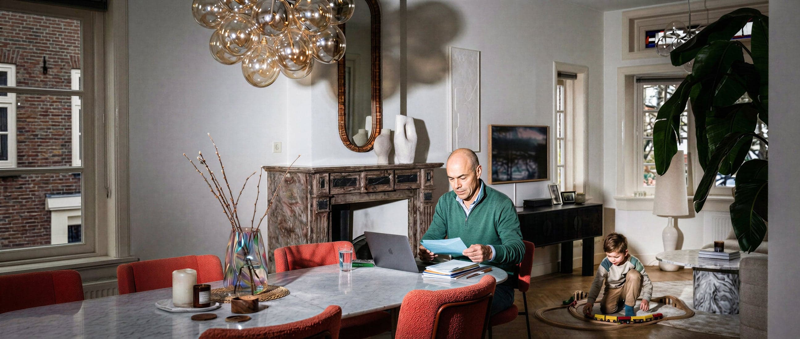 Persoon bekijkt documenten achter een laptop aan de eettafel, terwijl een kind speelt op een speelkleed in een stijlvolle woonkamer