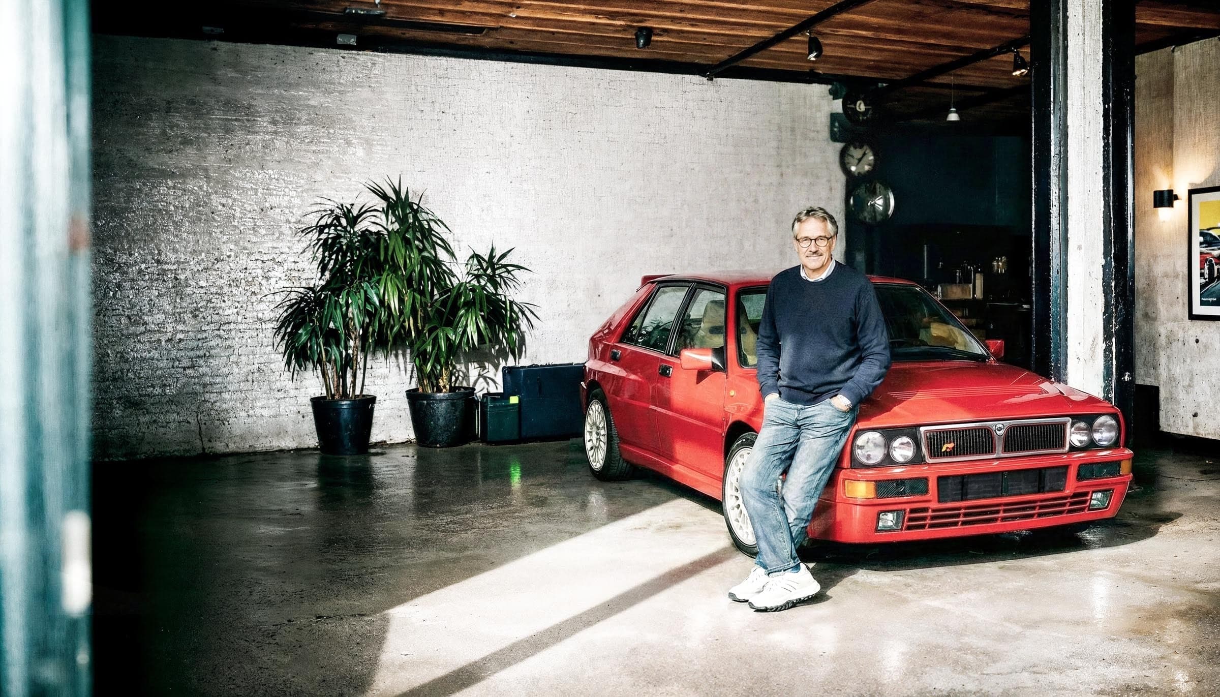Persoon leunt tegen een klassieke rode auto in een industriële ruimte met planten en een showroomachtige inrichting