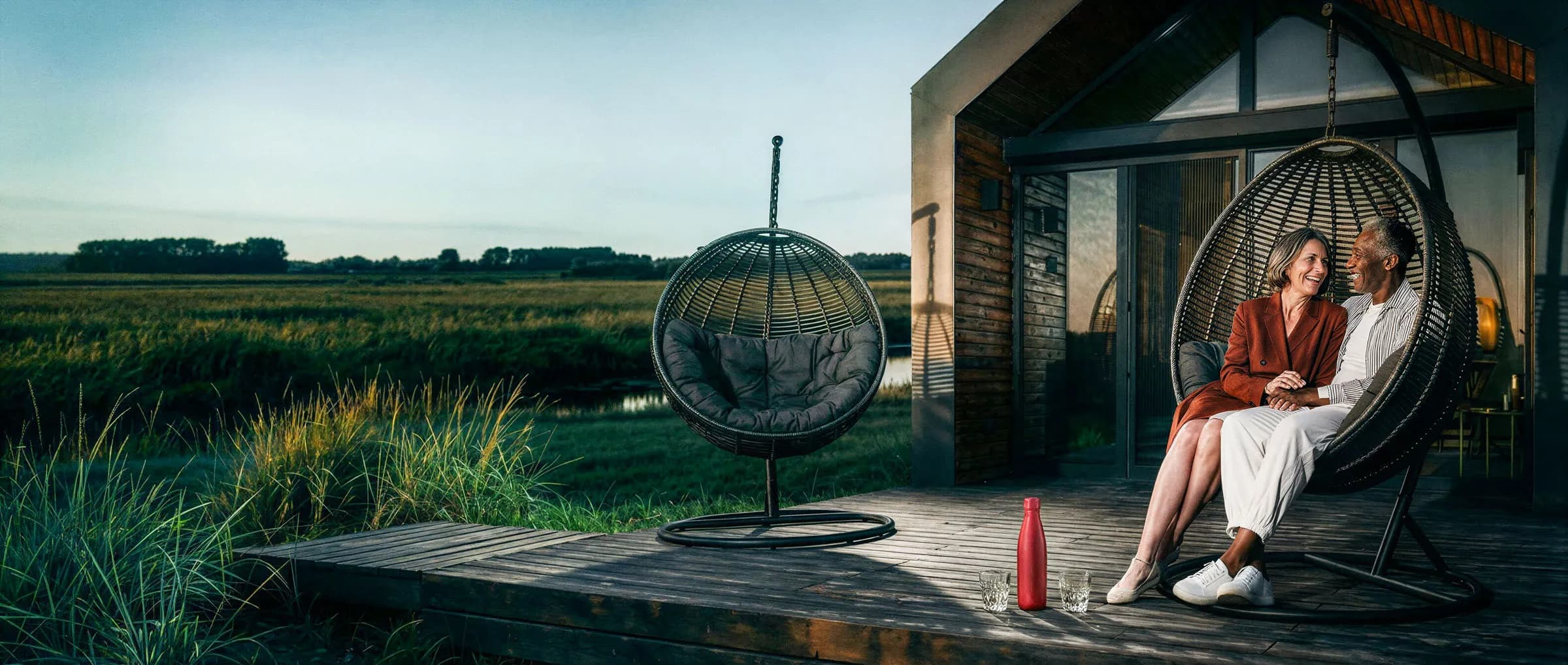 Twee personen zitten in een hangstoel op een houten terras naast een vakantiewoning, met uitzicht over een groen landschap