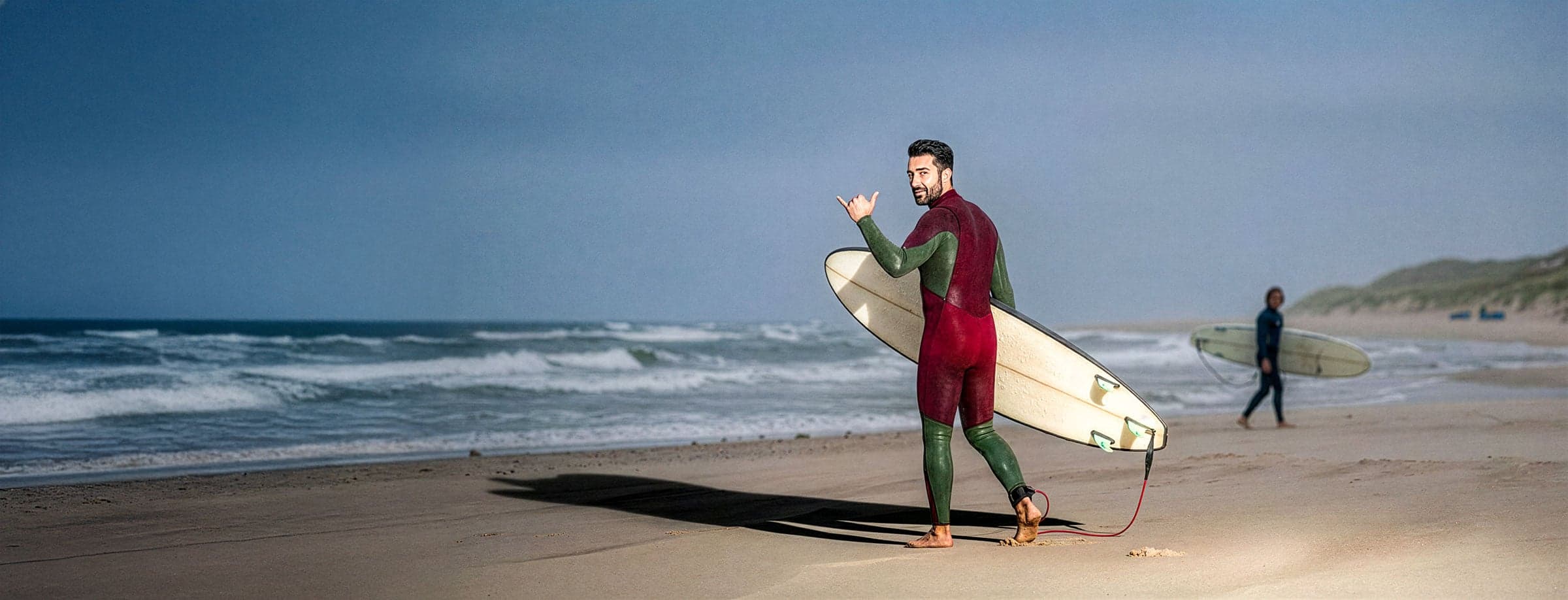 Surfer draagt een surfboard over het strand langs de branding, met een tweede surfer op de achtergrond