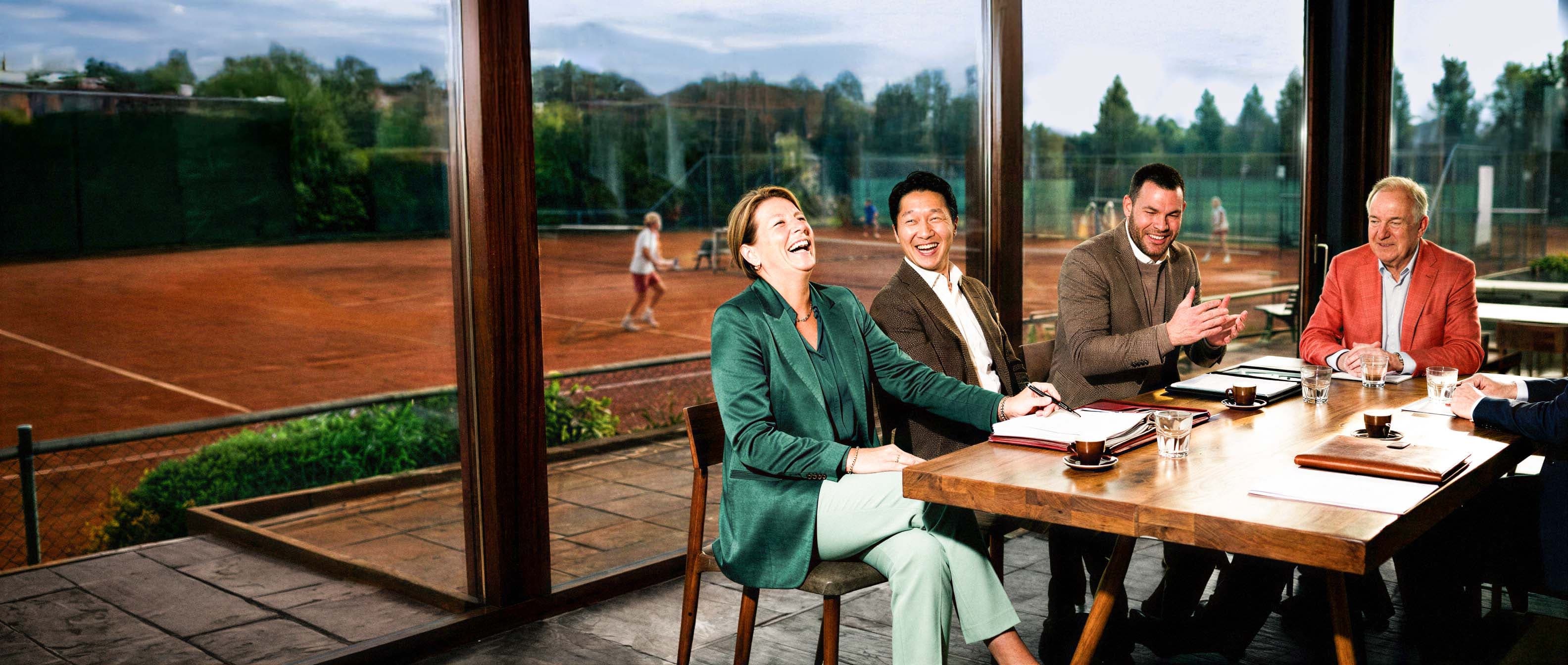Groep personen voert overleg aan een vergadertafel, met tennisbanen zichtbaar door de ramen op de achtergrond