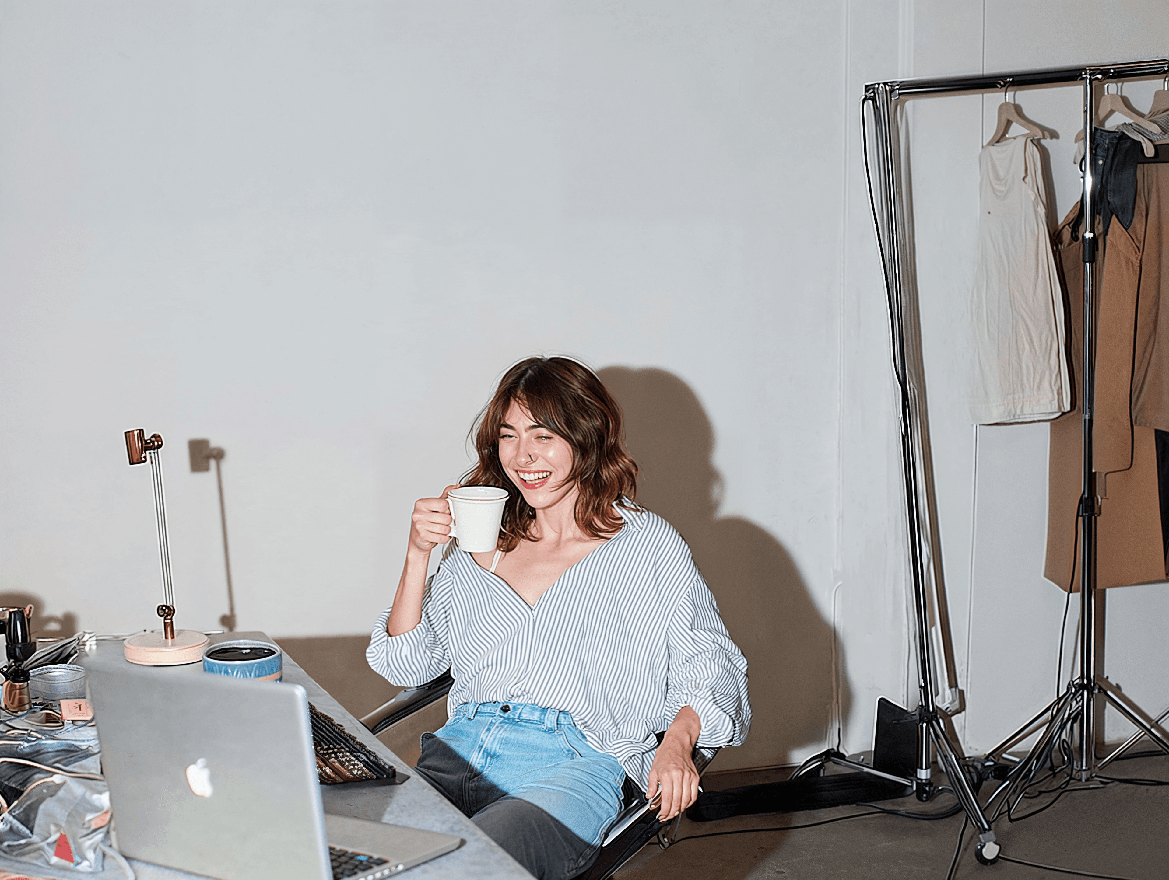 Persoon zit met een kop koffie achter een bureau met een laptop, terwijl kleding en accessoires in een studioomgeving zichtbaar zijn