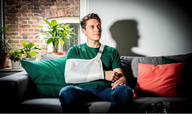 Persoon zit thuis op een bank met een arm in een mitella na een blessure of ongeval